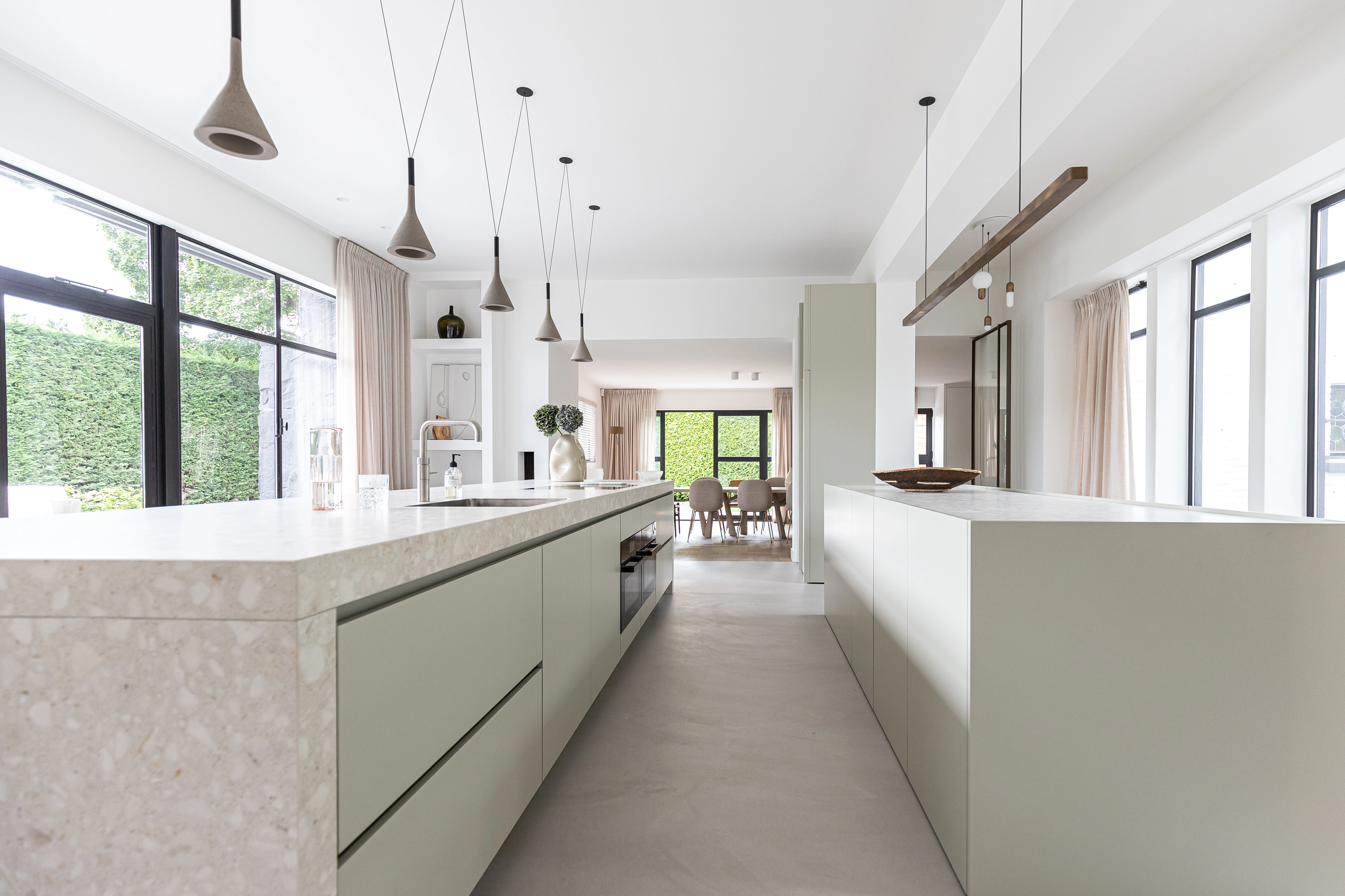 Lange open keuken met een terrazzo kookeiland, betonnen hanglampen en zwarte stalen kozijnen met uitzicht op de tuin