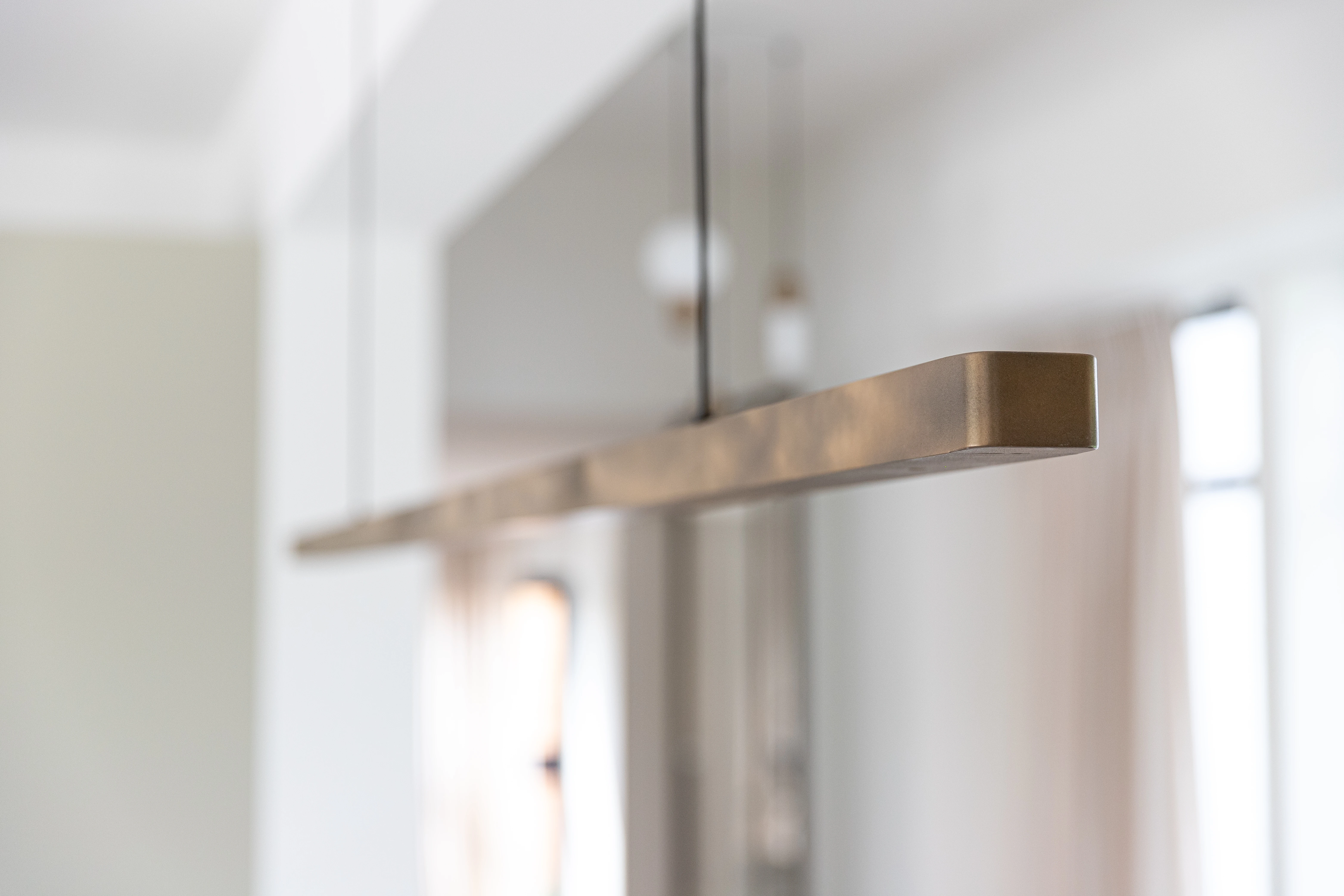 Detail van een langwerpige hanglamp in brons voor een raam met gordijn