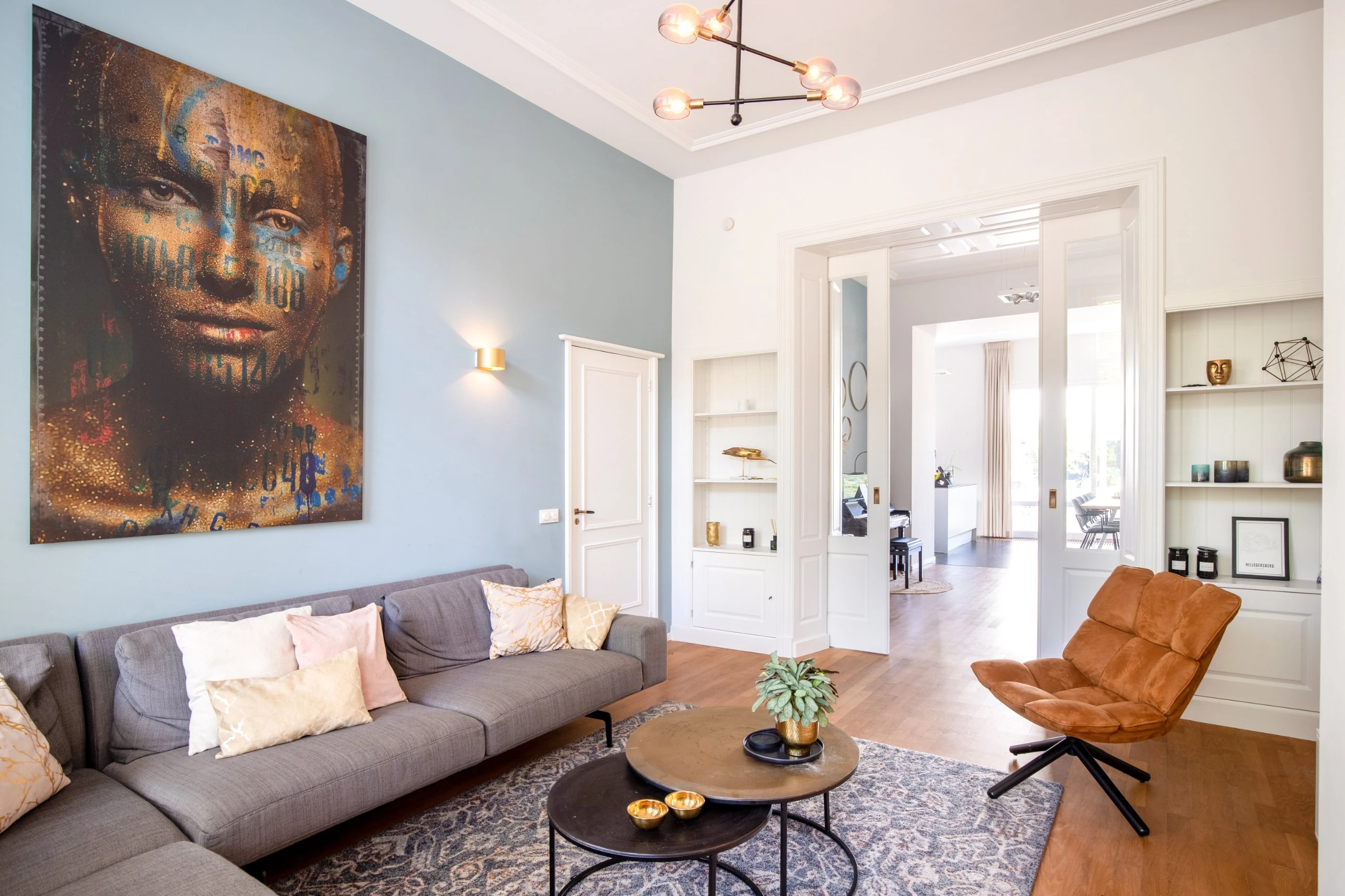 Woonkamer met een lichtblauwe wand, een groot kunstwerk, een grijze hoekbank en schuifdeuren met doorkijk naar de keuken