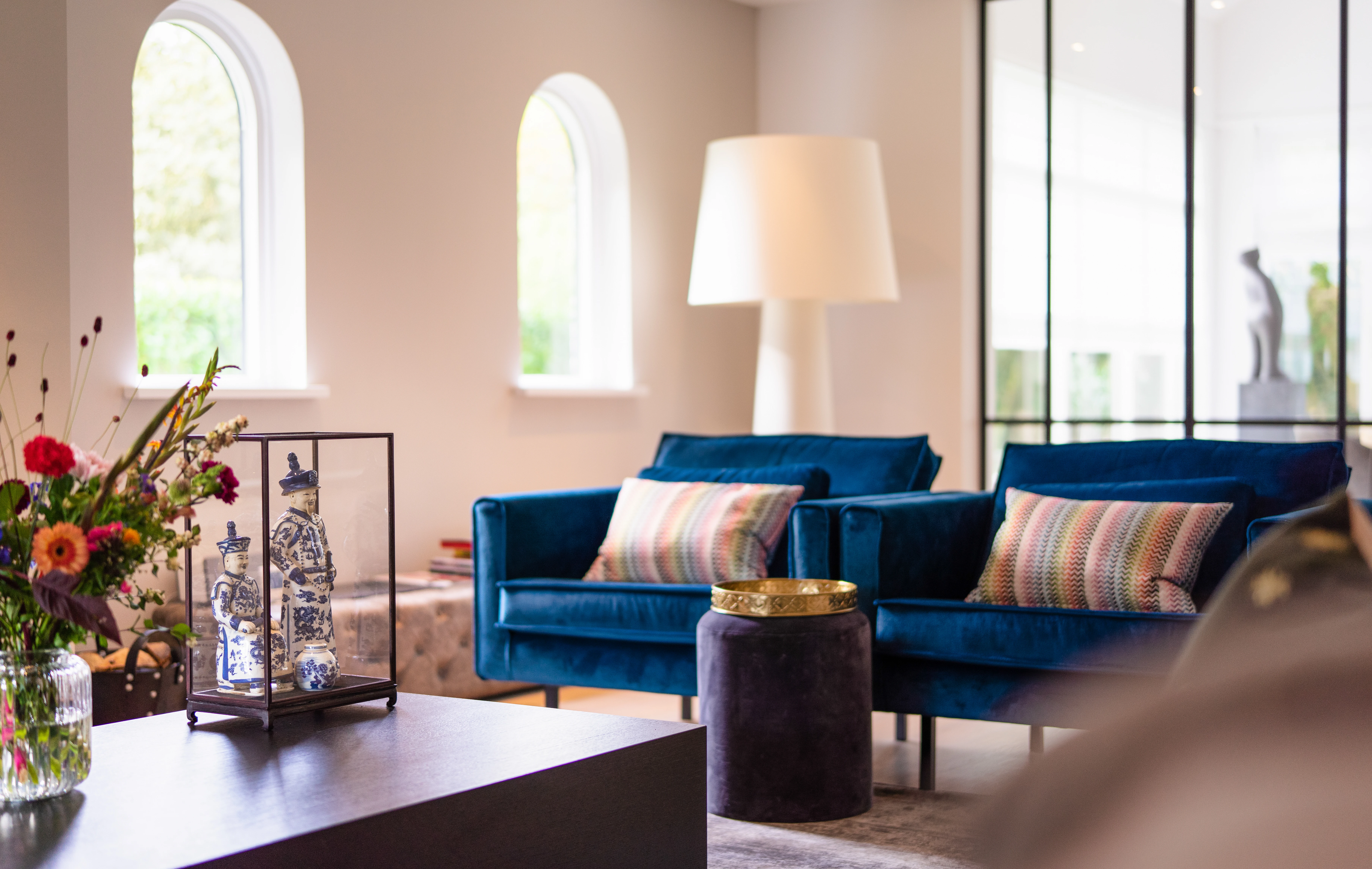 Hoek van de woonkamer met boogramen, twee blauwe fauteuils en een vitrine met porseleinen beeldjes