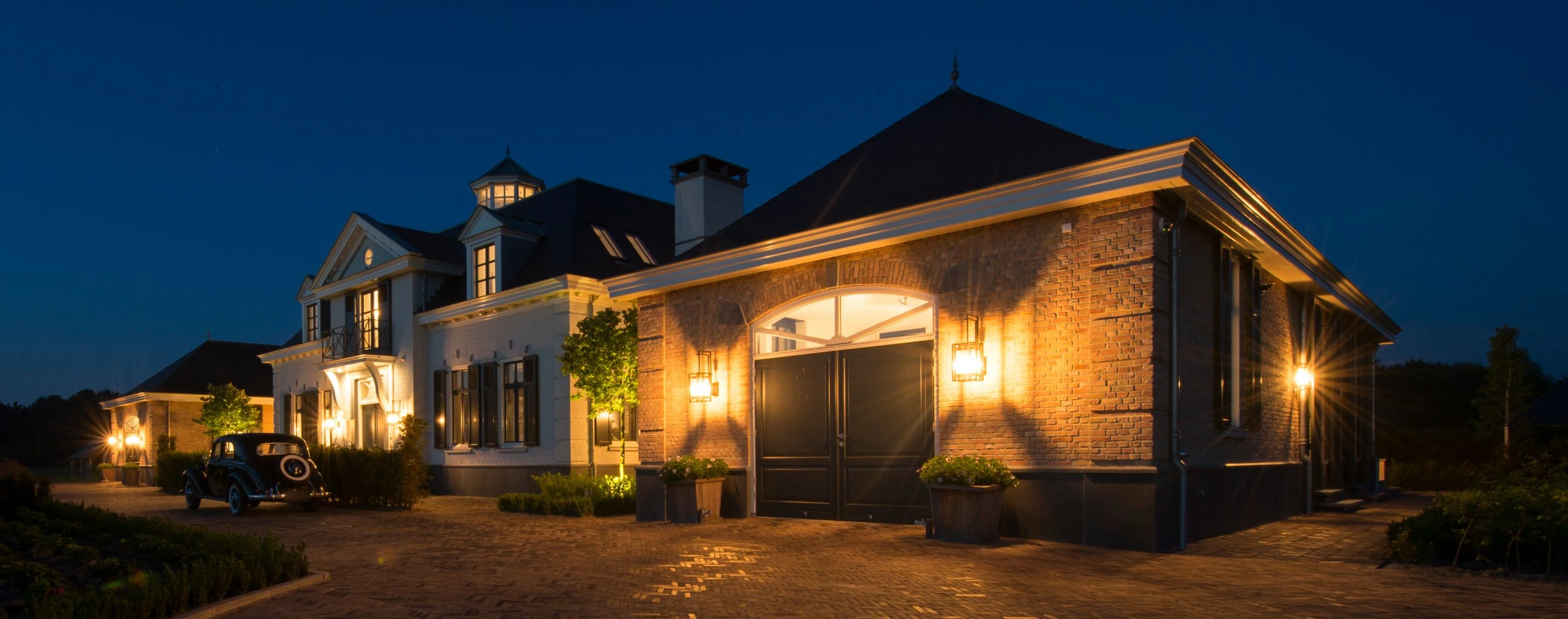 Villa in Sommelsdijk bij avondlicht, met verlichte gevel, een garage met boograam en een oldtimer op de oprit