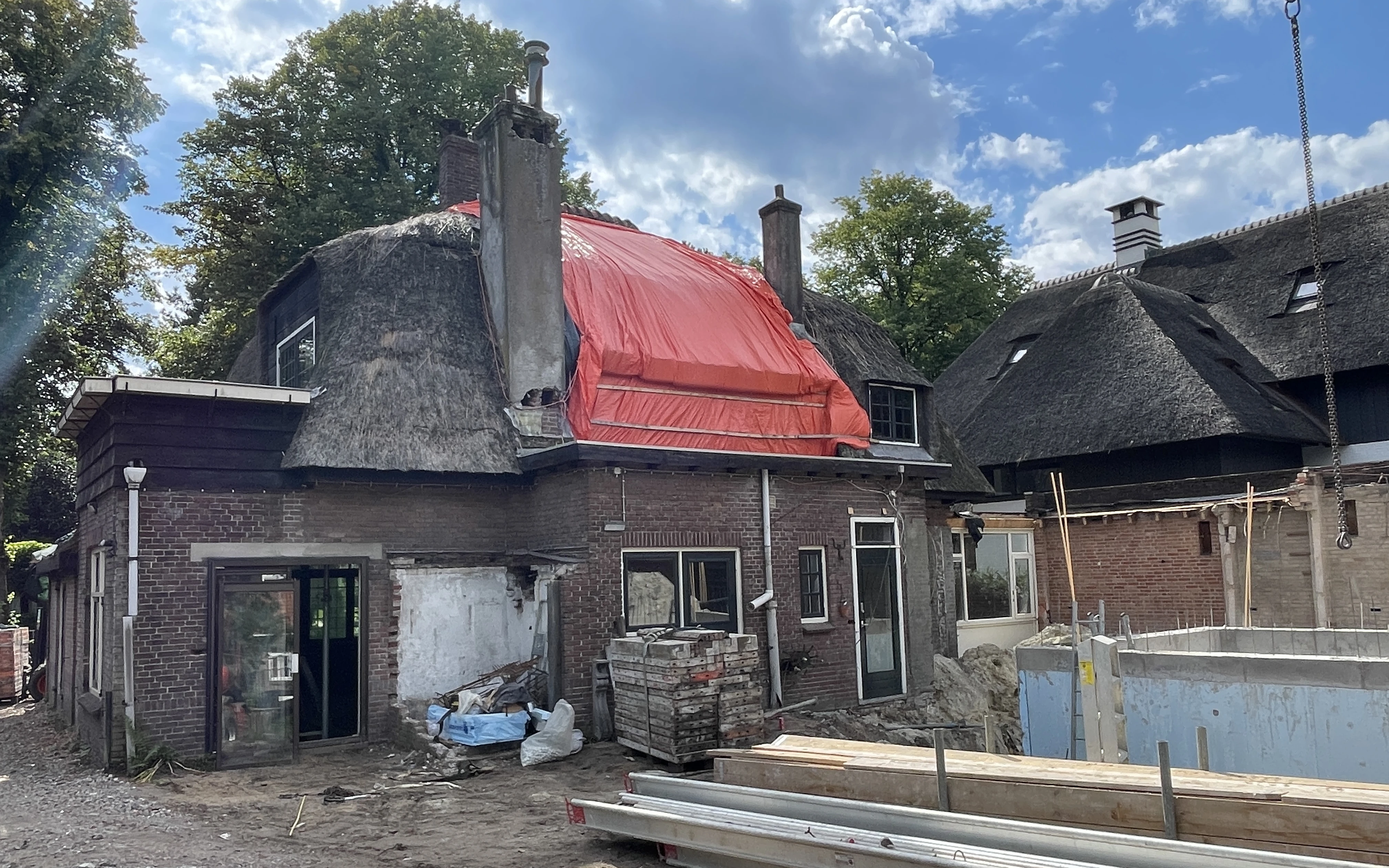 Achterzijde van de rietgedekte villa tijdens de verbouwing, met een rood dekzeil over de kap en gestript metselwerk