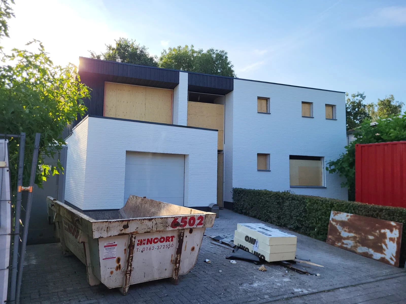 Voorgevel van de woning tijdens de verbouwing, met deels dichtgetimmerde kozijnen en een afvalcontainer op de oprit