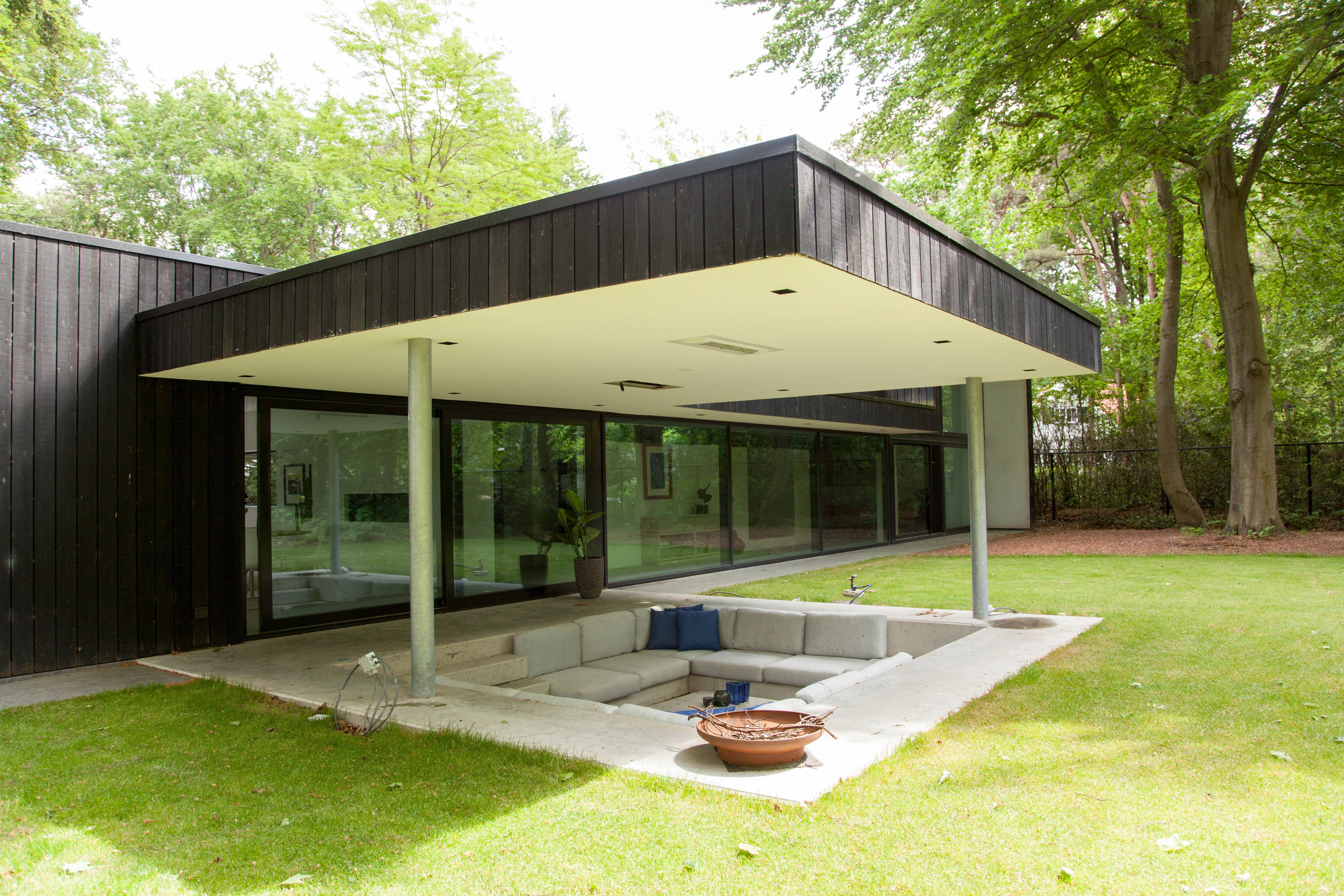 Overkapt terras met een verzonken loungehoek bij een villa met zwart gevelhout, gezien vanaf het gazon